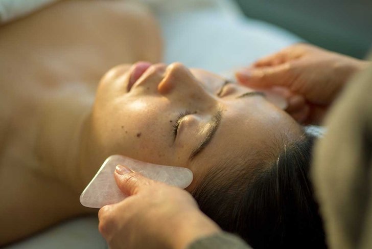 Young woman receiving facial rejuvenation gua sha with rose quartz in acupuncture wellness spa clinic