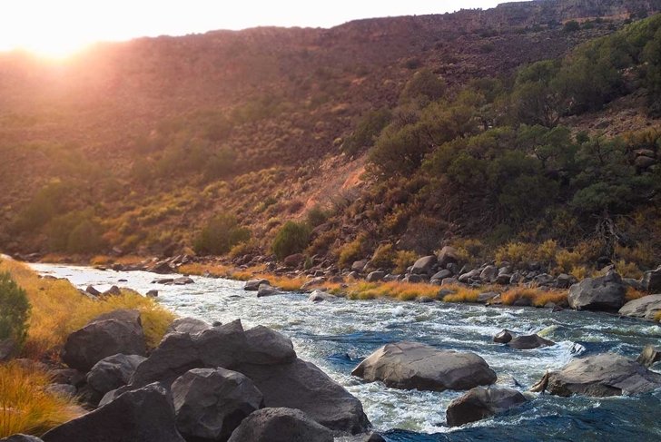 Riverside view of the Rio Grande River in New Mexico. The sun dips is just about to dip below the gorge rim as the river babbles across rocks in the forefront.