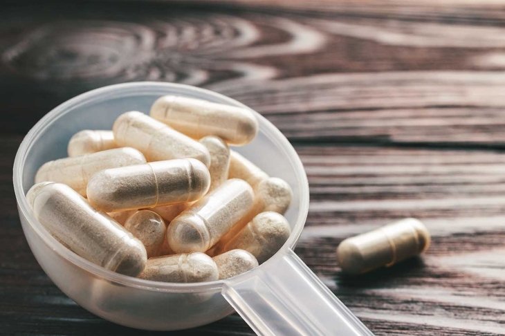 White medical capsules of glucosamine chondroitin, healthy supplement pills in the plastic spoon on wooden background, macro image.