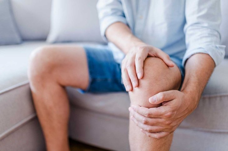 Man suffering from knee pain sitting sofa. A mature man massaging his painful knee. Man suffering from knee pain at home, closeup. Pain knee