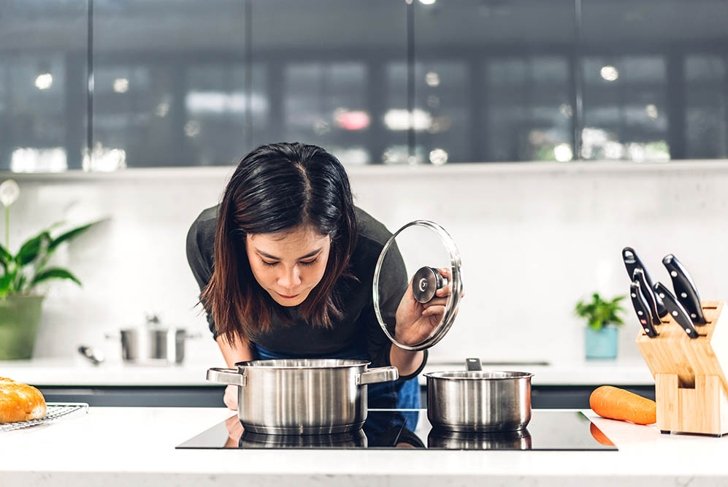 Young asian woman standing near stove and cooking.Happy woman looking and smelling tasting fresh delicious from soup in a pot with steam at white interior kitchen